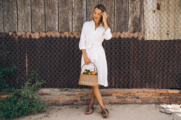 picture of a woman in white summer dress and slip on sandals holding a basket of orange standing in front a cottage