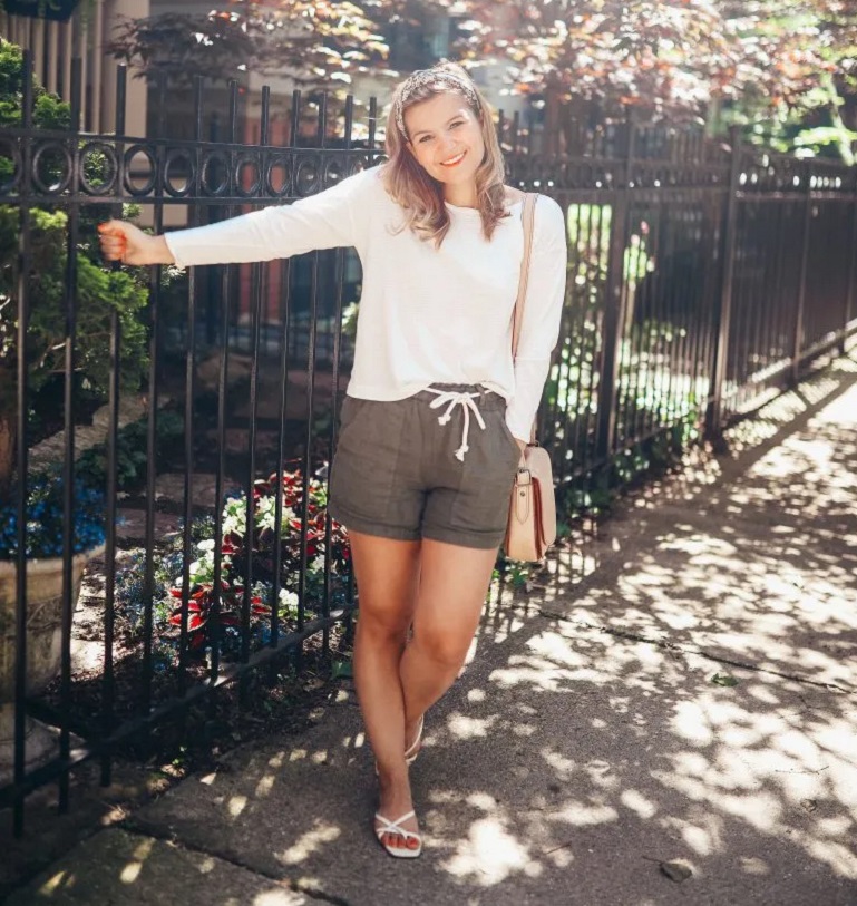 picture of a woman on a sidewalk wearing long sleeves top, linen paper bag shorts and slip on sandals 